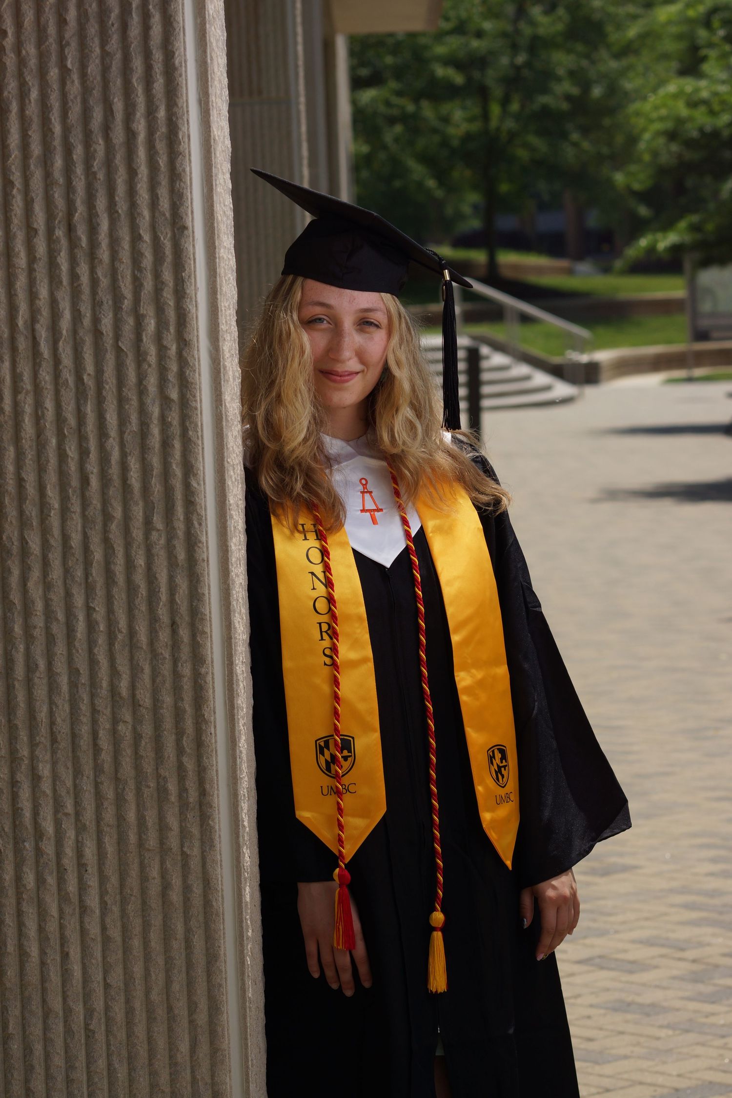 Smiling female graduate in black cap and gown with gold honor stole posing outdoors on college campus.