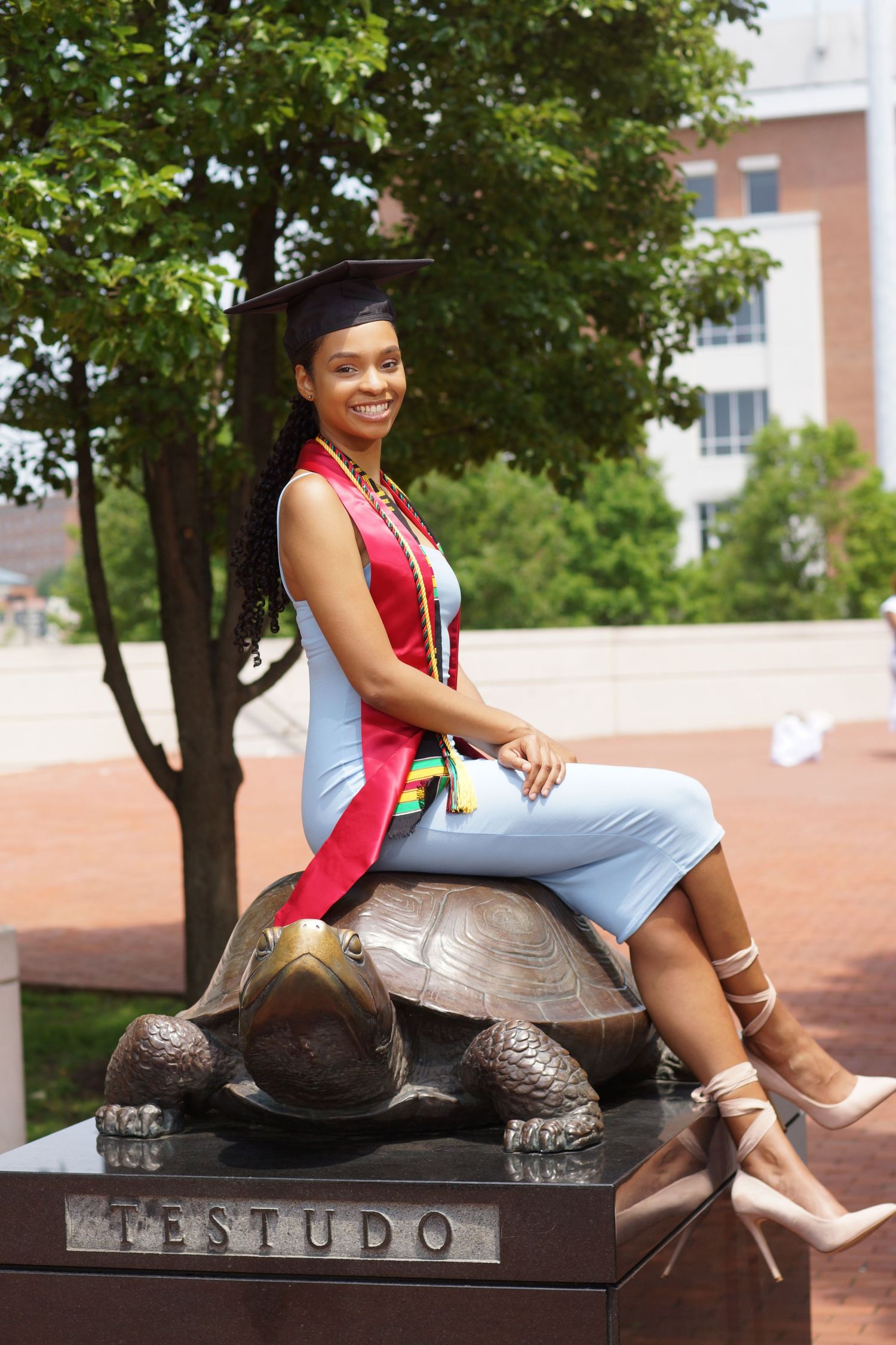 A smiling graduate in cap and gown sits atop the Testudo bronze turtle statue at the University of Maryland.