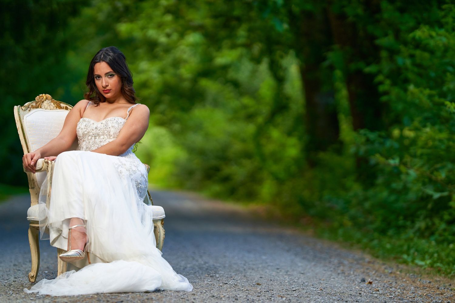 A person in a flowing white wedding dress sits gracefully on an elegant cream-colored chair on a forest path.