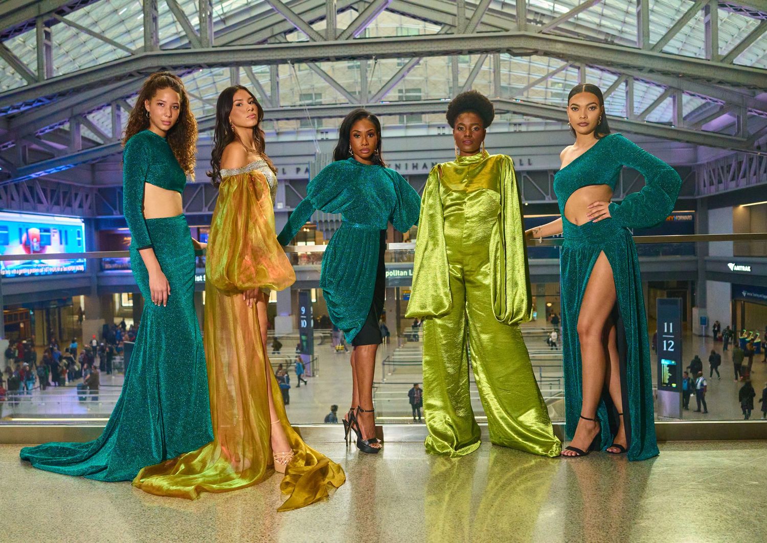 Fashion models in elegant jewel-toned dresses pose together in industrial venue setting.