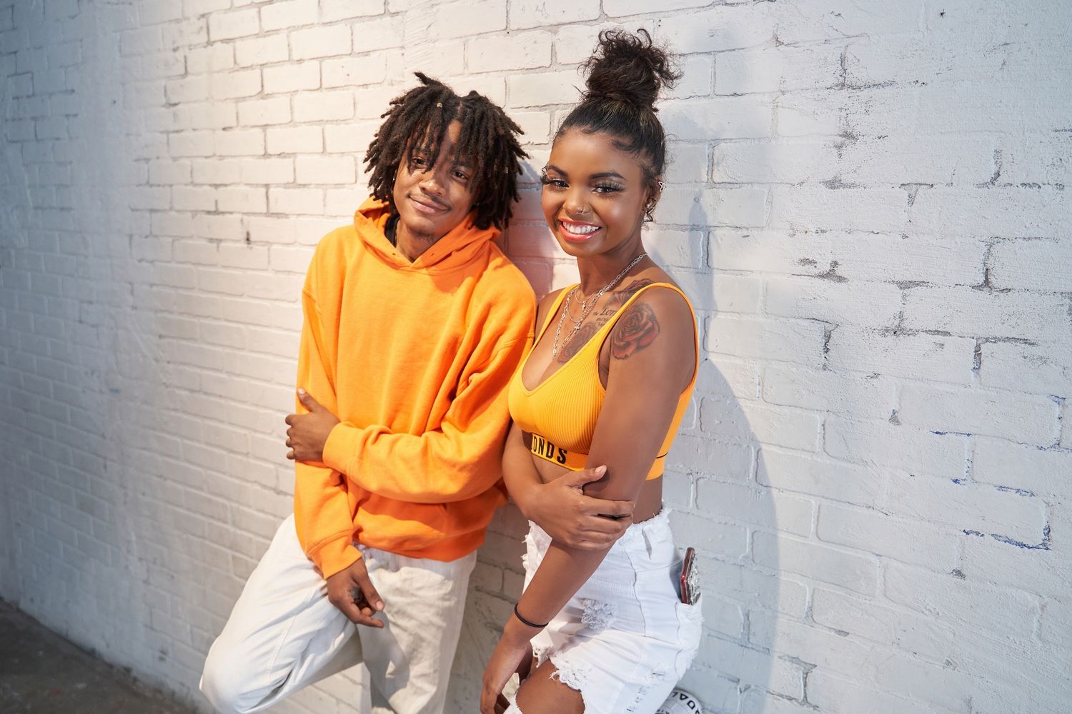 Fashion models posing playfully against white brick wall in orange and white outfits.