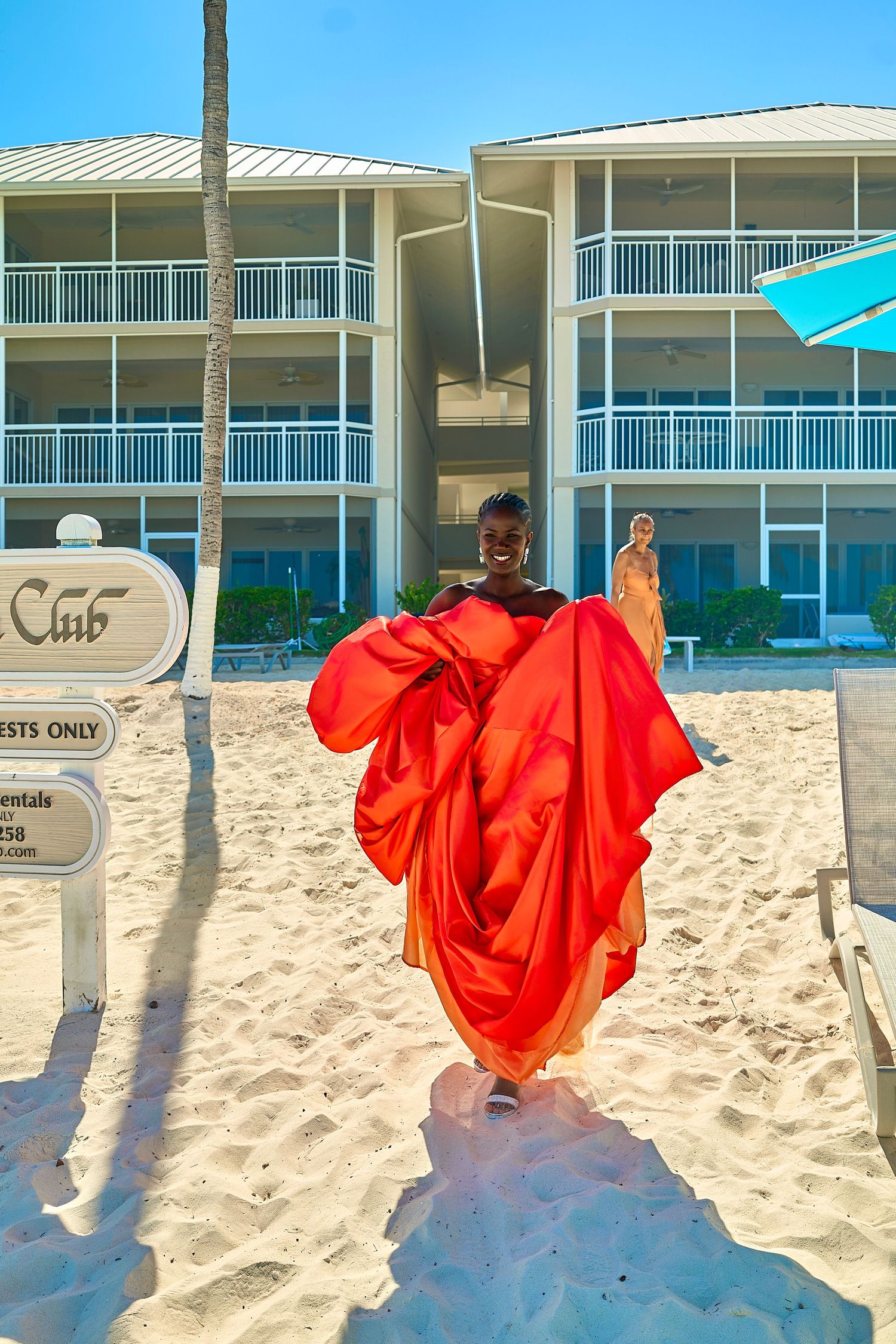 Woman in flowing red dress walking on white sand beach in front of a resort building on a sunny day.