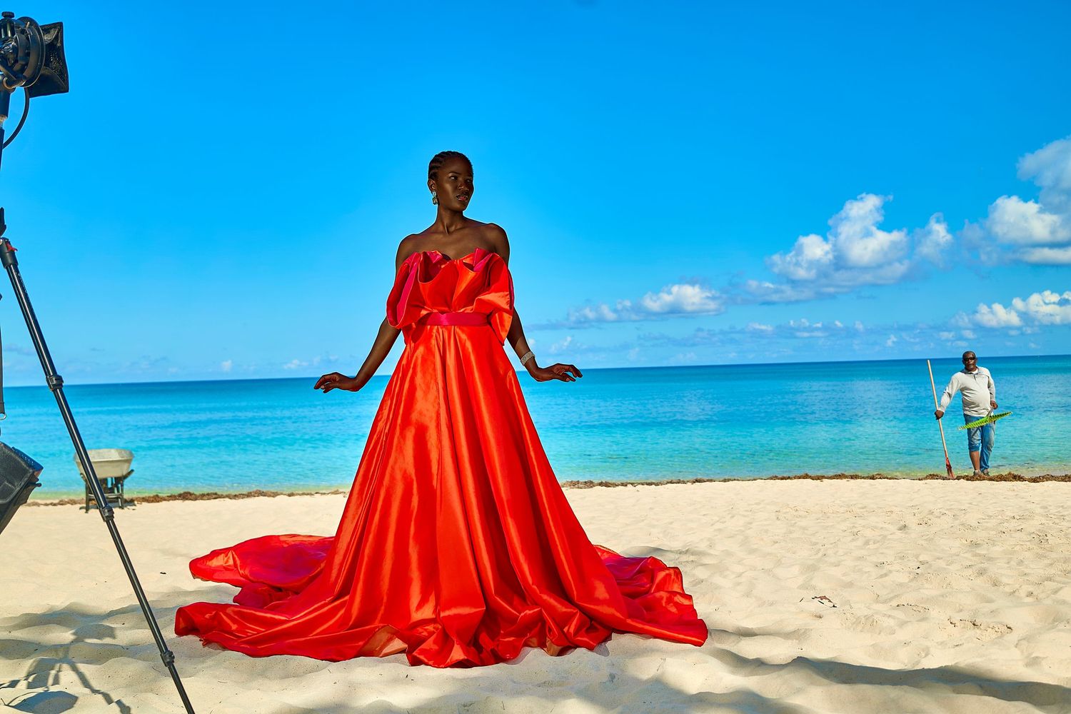 A woman in a stunning red ballgown poses on a tropical beach with clear blue skies and ocean in the background.