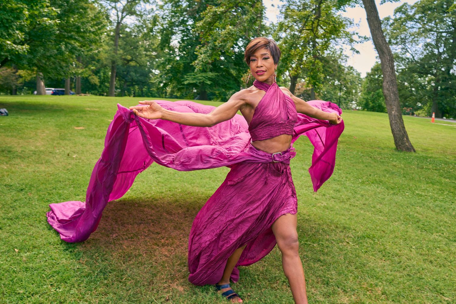 Woman in flowing pink outfit dancing gracefully in a lush green park, performing an elegant belly dance routine.
