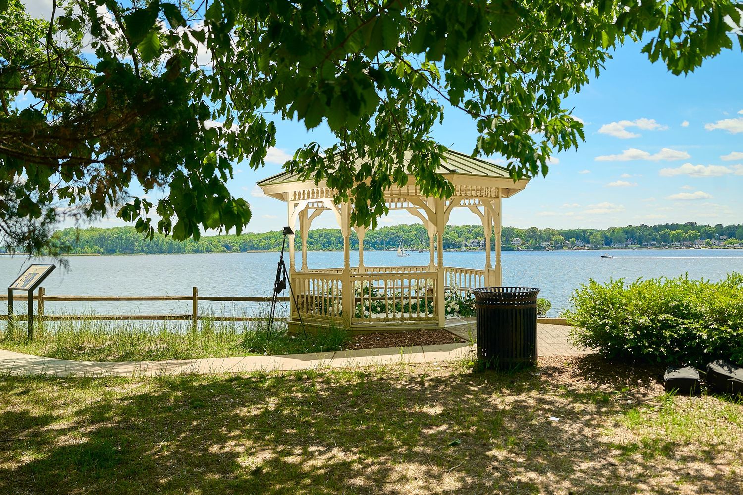 White gazebo by a serene lakeside park with lush green trees and a trash bin on a sunny day.