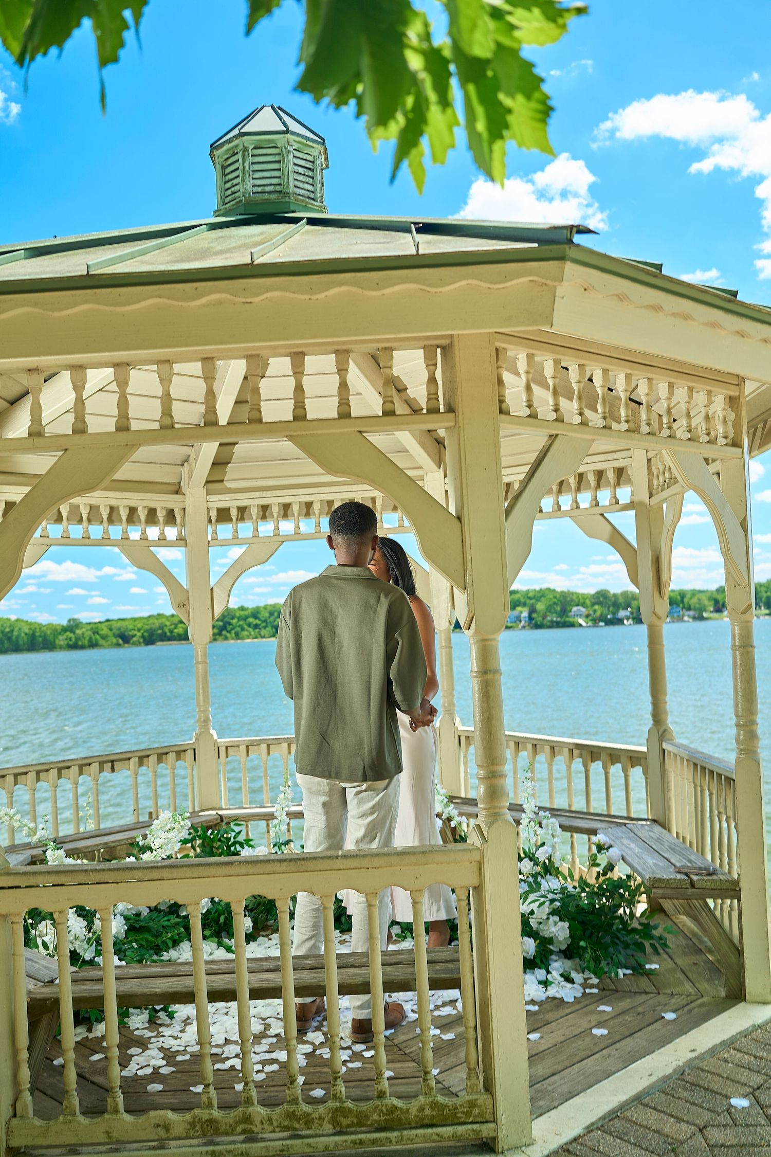 Couple standing together in a white lakeside gazebo decorated with white flowers on a sunny day.
