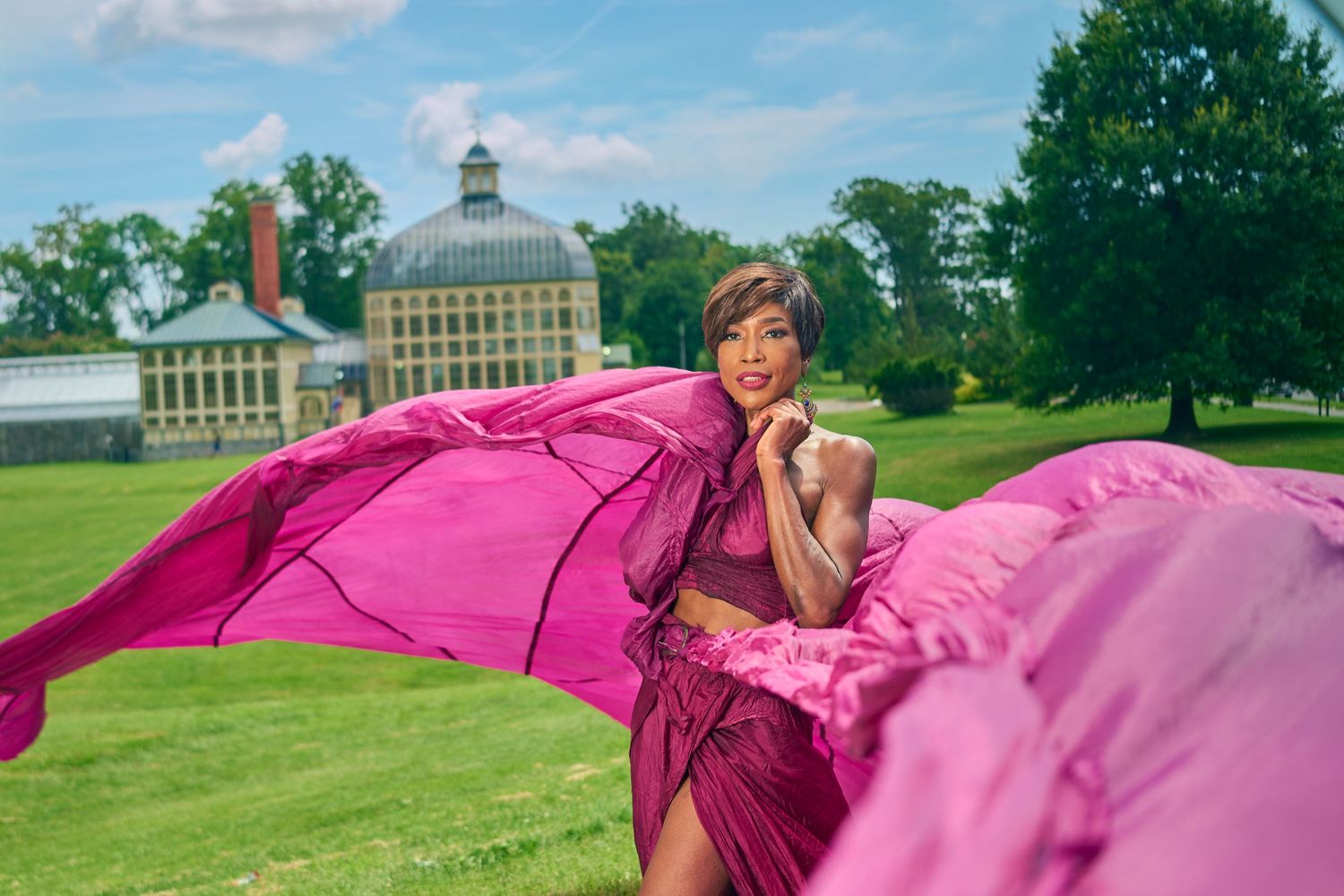 Woman in flowing pink gown posing outdoors on green lawn with historic building backdrop on sunny day.