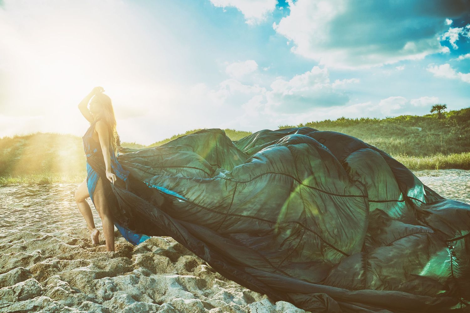 Teal fabric billows ethereally against sunset sky on beach landscape.