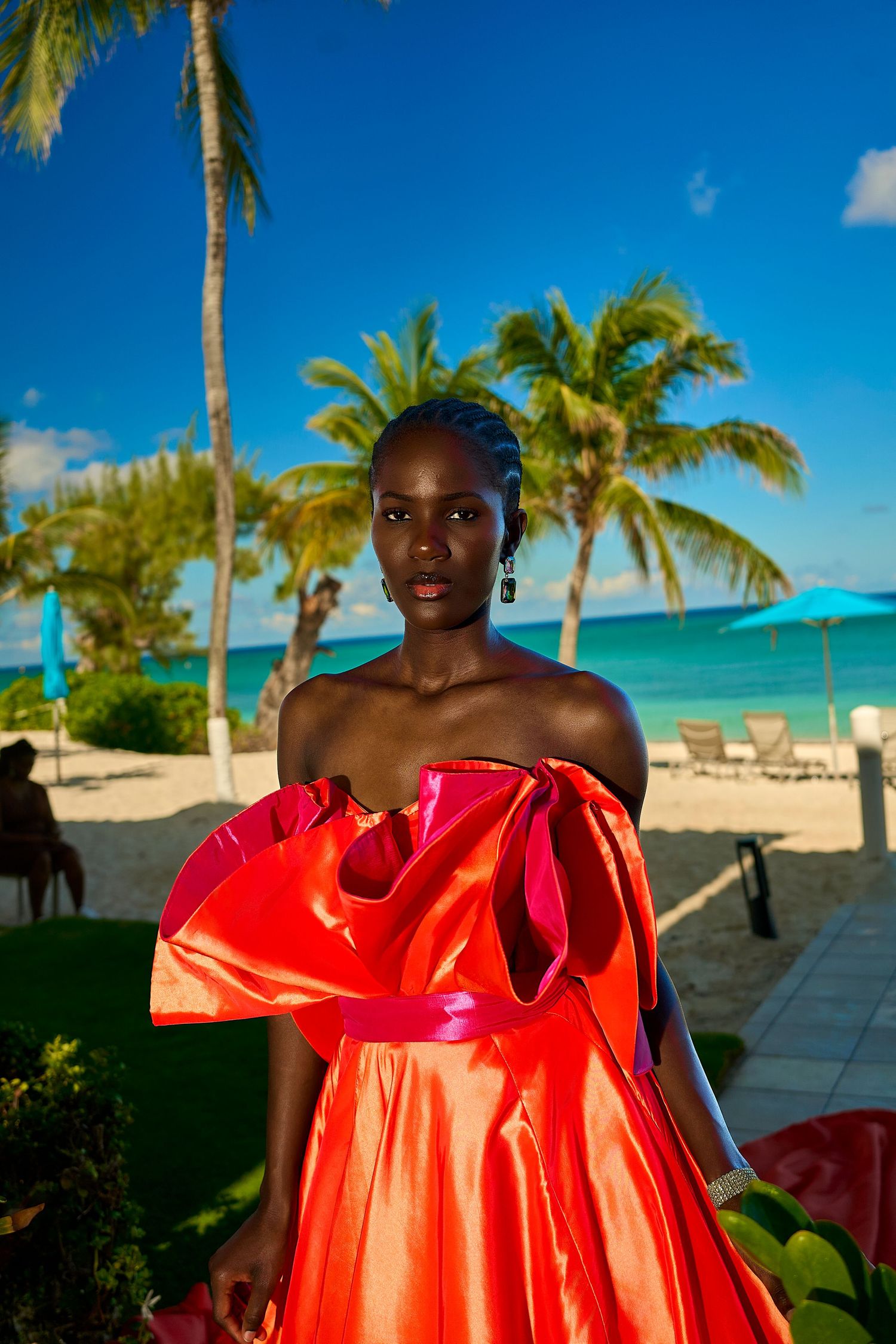 A Black woman in a stunning off-shoulder orange satin gown poses on a tropical beach with palm trees and turquoise water.
