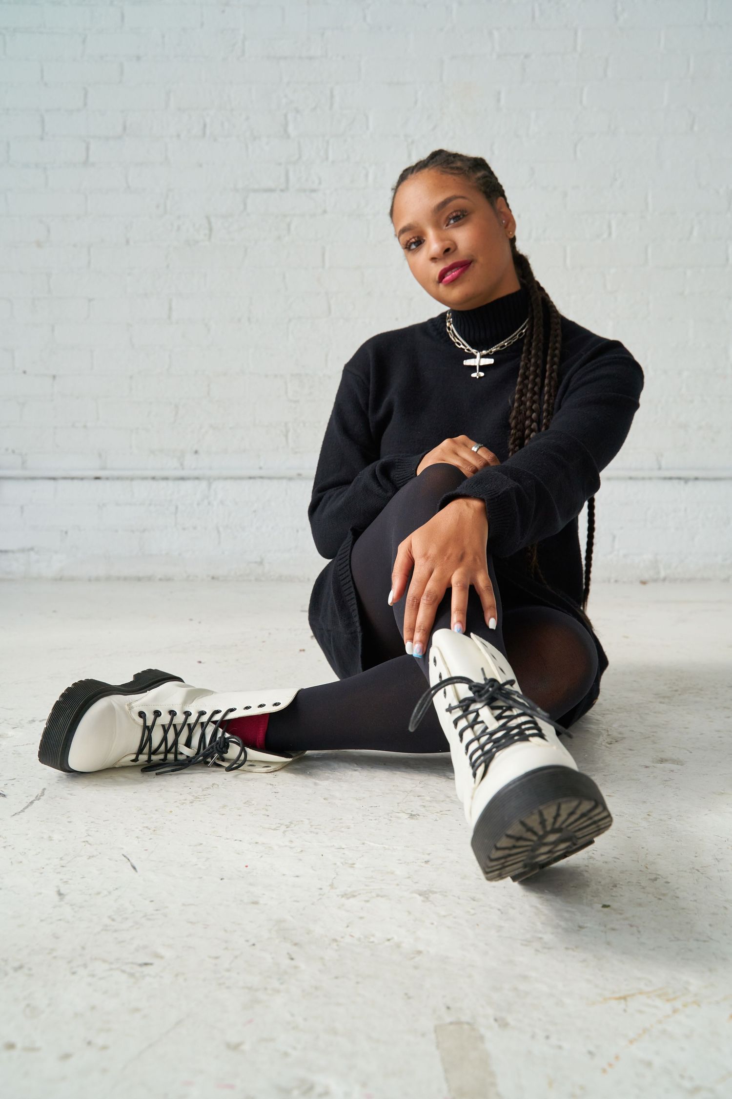 Person in black clothing sitting on floor showing off black and white sneakers against white wall.