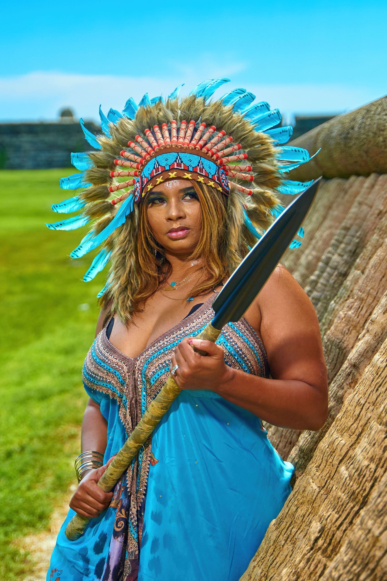 Person wearing turquoise dress and feathered headdress poses against natural outdoor backdrop.