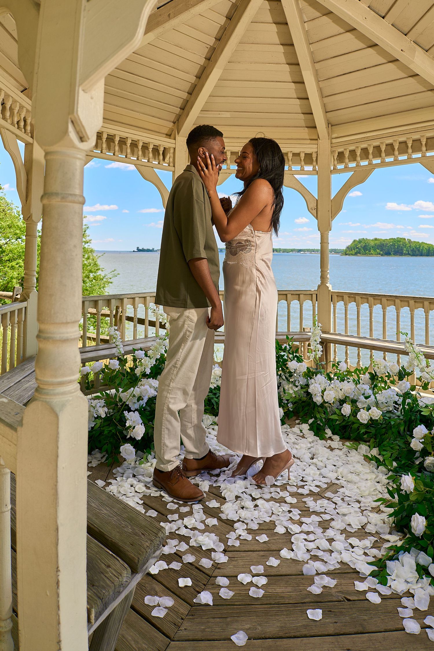 Couple shares a kiss in a flower-adorned gazebo with white rose petals and lake views.
