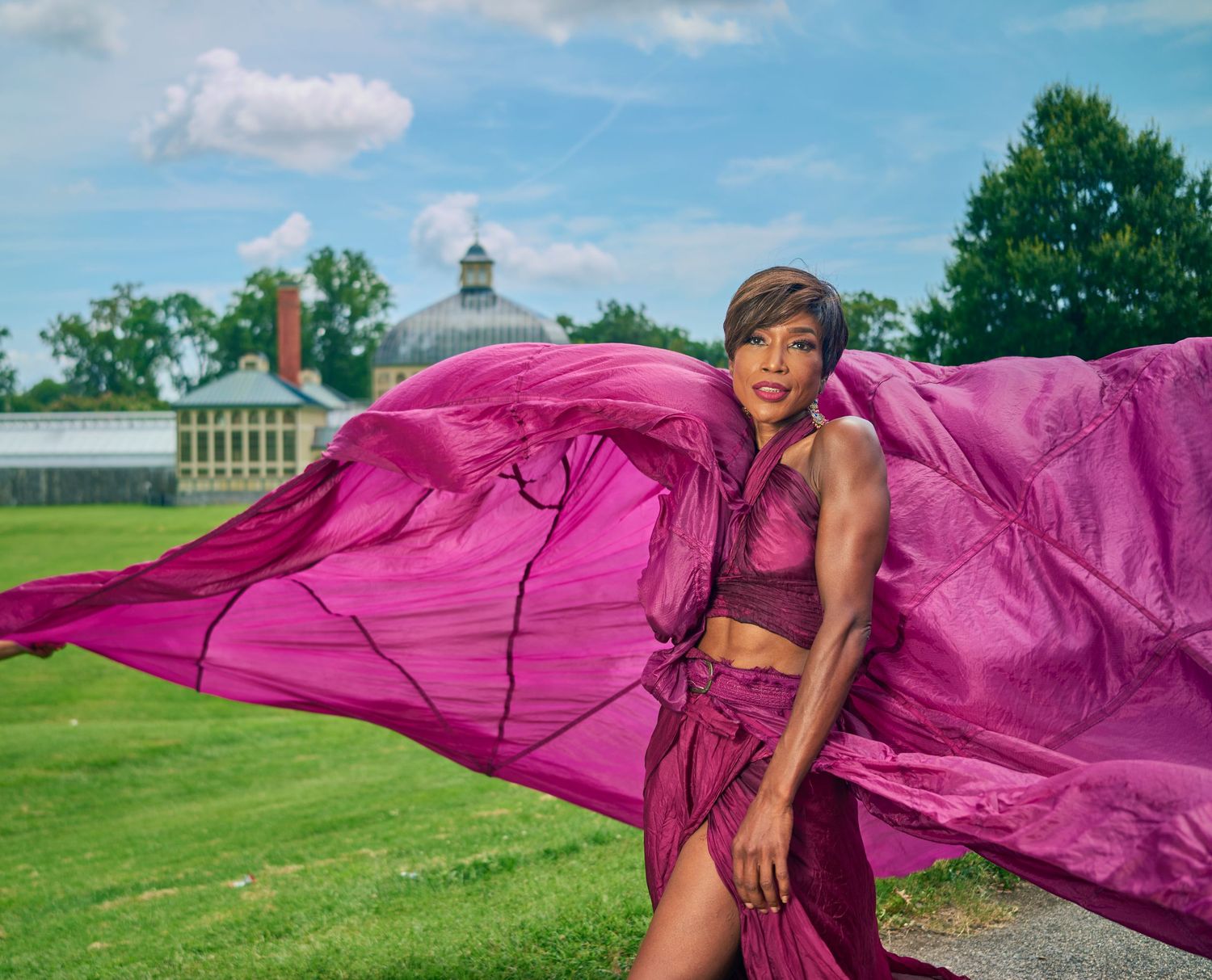 Elegant woman in magenta two-piece outfit with billowing fabric poses on lawn near historic greenhouse.