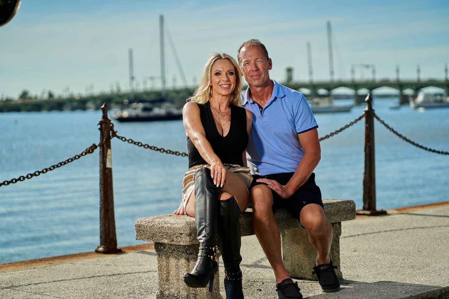 Couple poses for romantic photos on a waterfront boardwalk with boats and a bridge in the background.