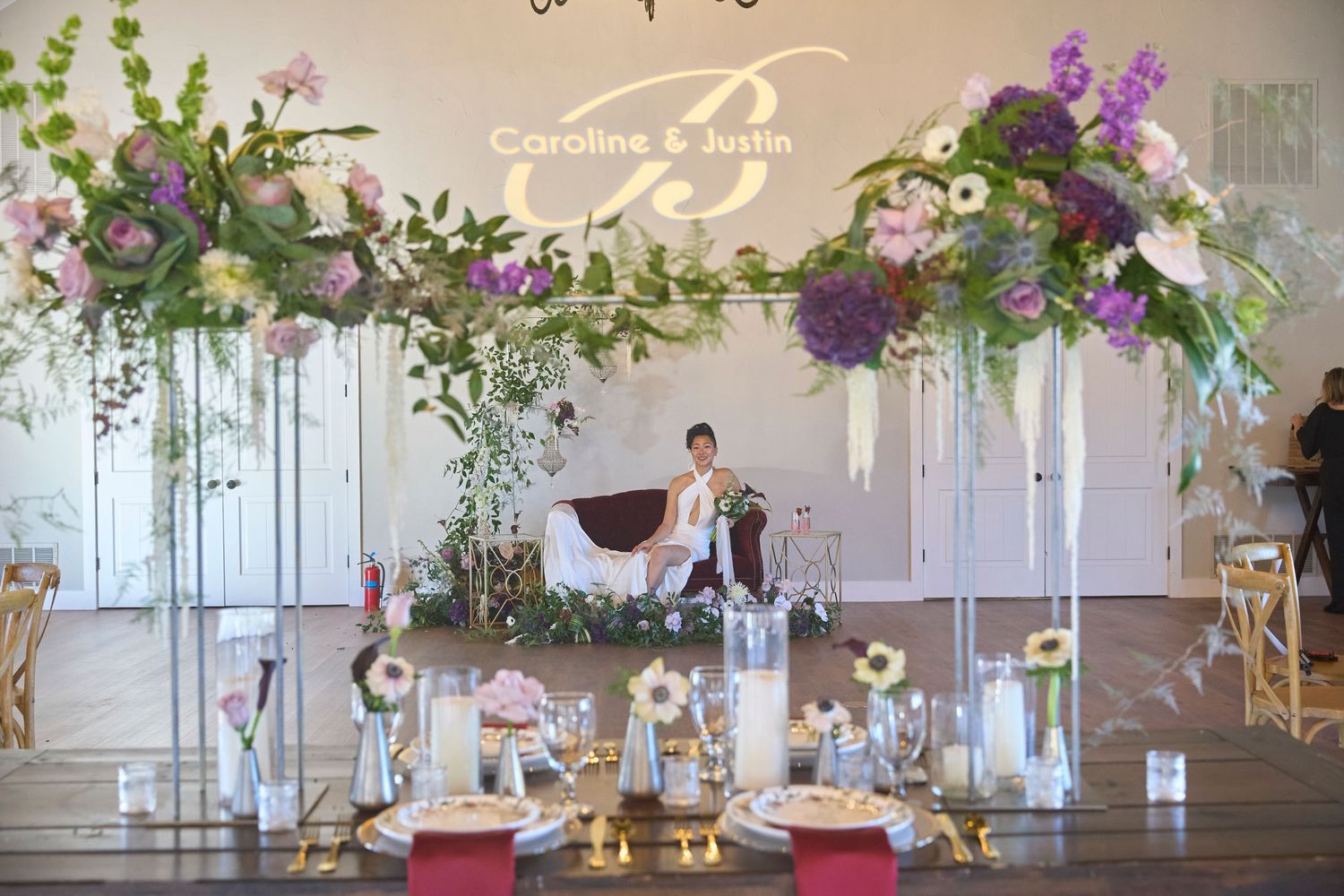 Modern wedding reception venue decorated with elegant purple floral arrangements and chandeliers.