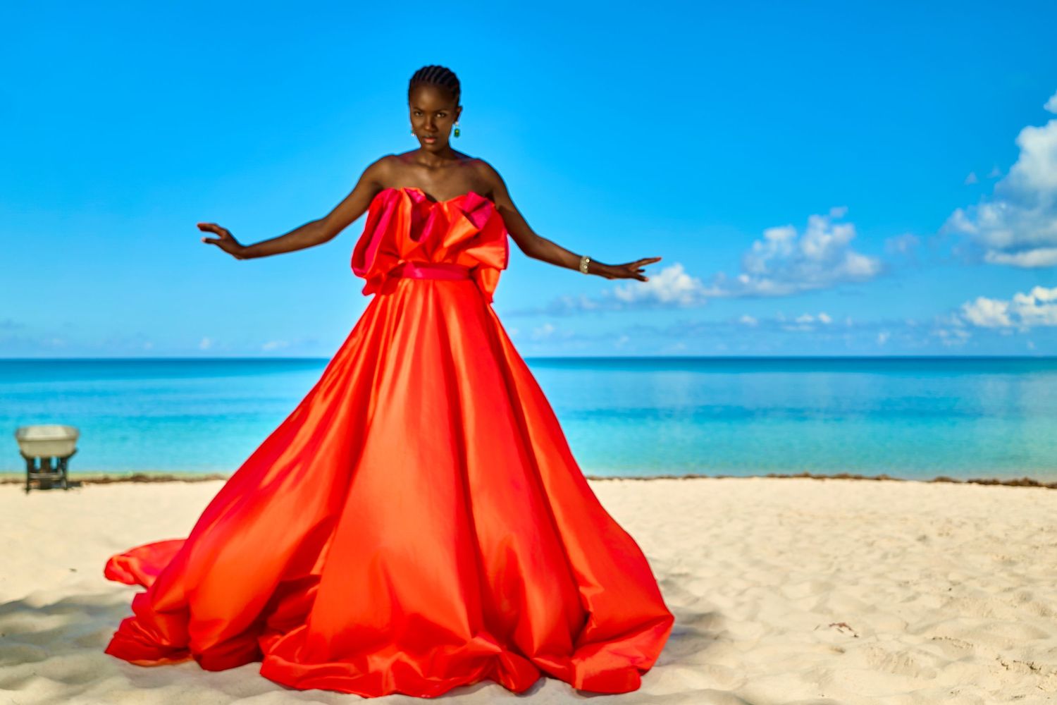 Woman in stunning red ball gown poses gracefully on a tropical white sand beach under a bright blue sky.