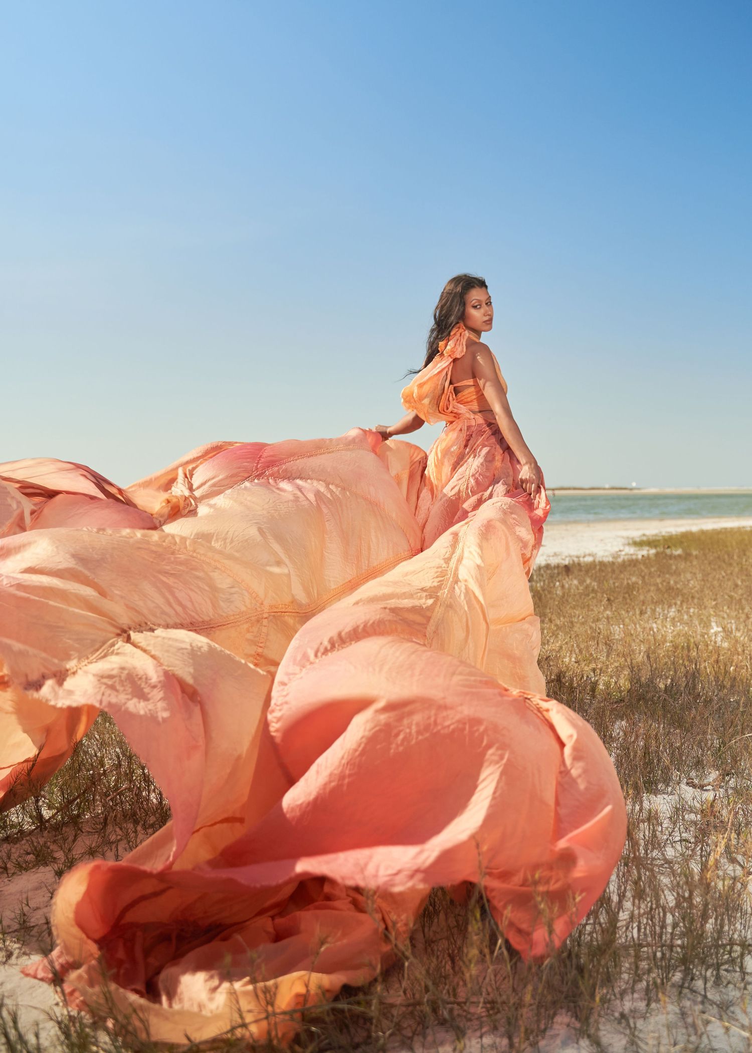 Flowing coral dress billows dramatically against beach grass and sand dunes.