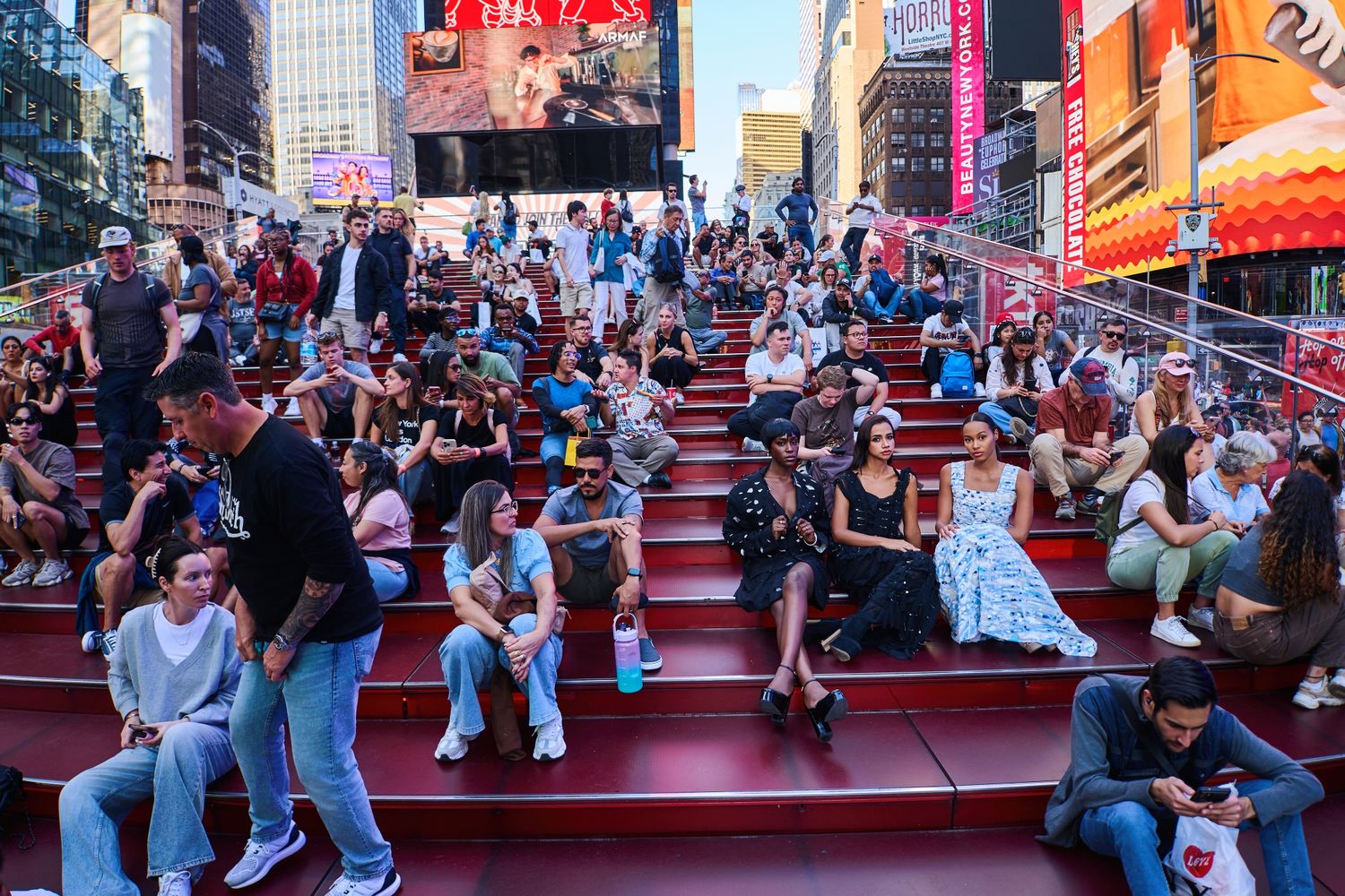 Crowded outdoor urban plaza with red steps filled with people sitting and socializing.