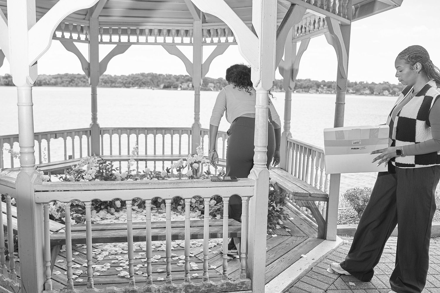 Two women near a white wooden gazebo overlooking a serene lake, one holding a box outdoors.