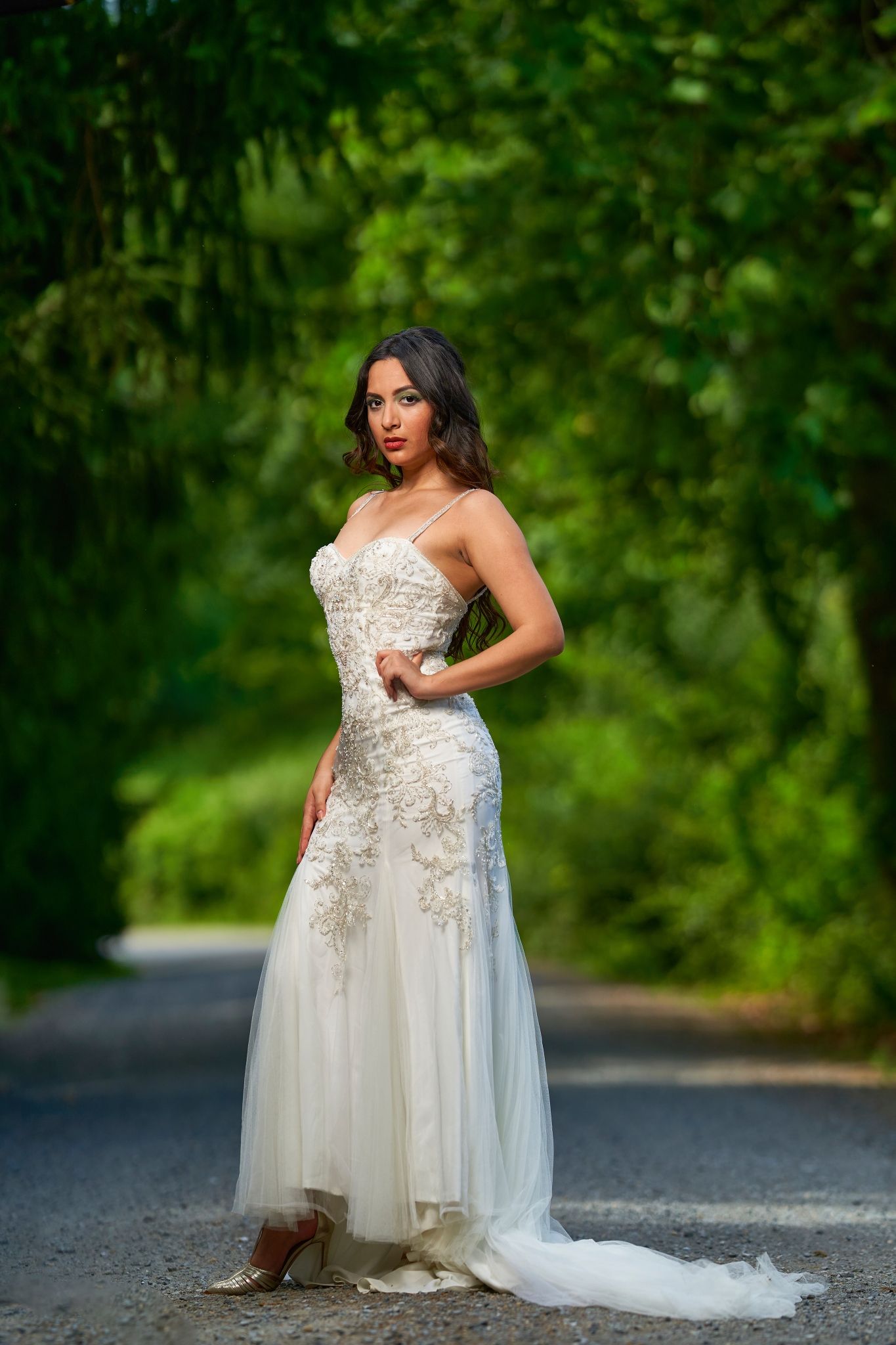 A bride in a flowing white lace gown with floral appliques poses outdoors against a lush green background.