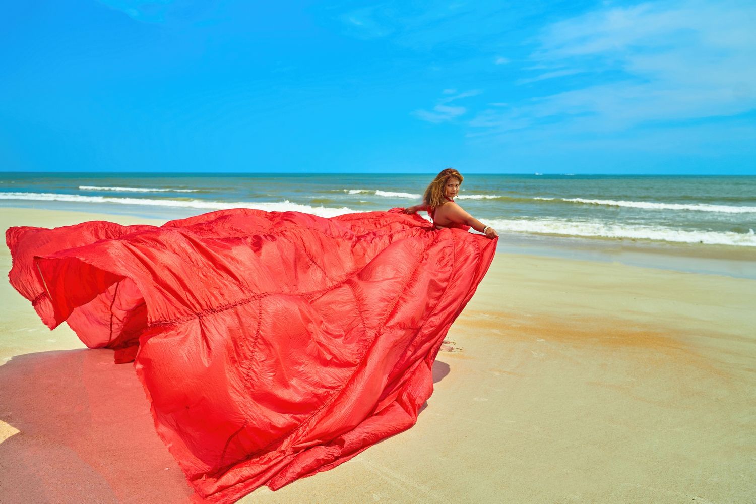 A flowing red dress catches the wind on a sunny beach against blue ocean waves.