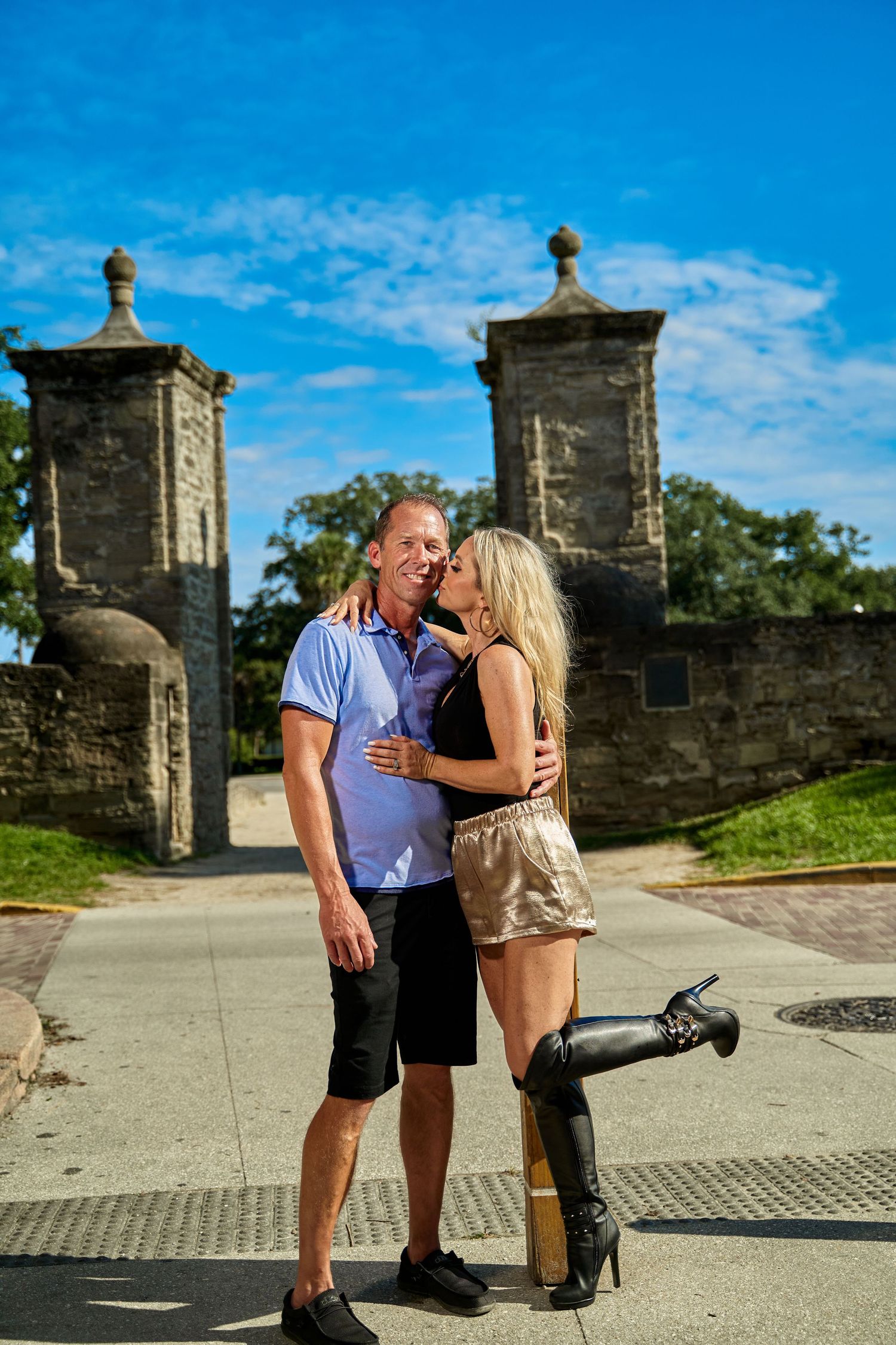 Couple sharing romantic moment near historic stone gate structure under blue sky.