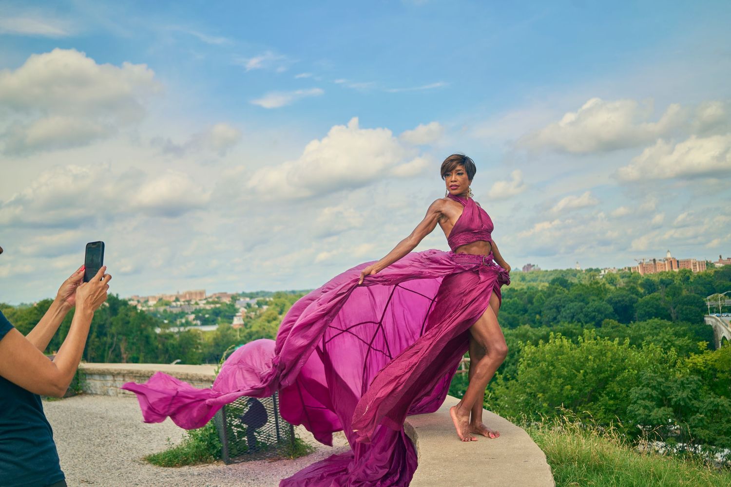 Photographer capturing woman in billowing magenta dress on scenic overlook hilltop.