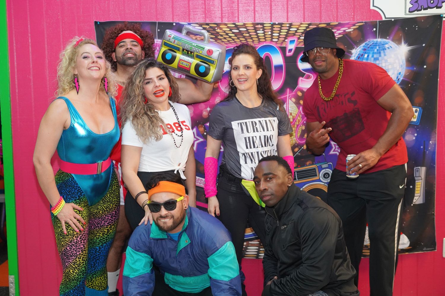 Group of dancers in colorful outfits performing energetic dance moves against a pink backdrop.