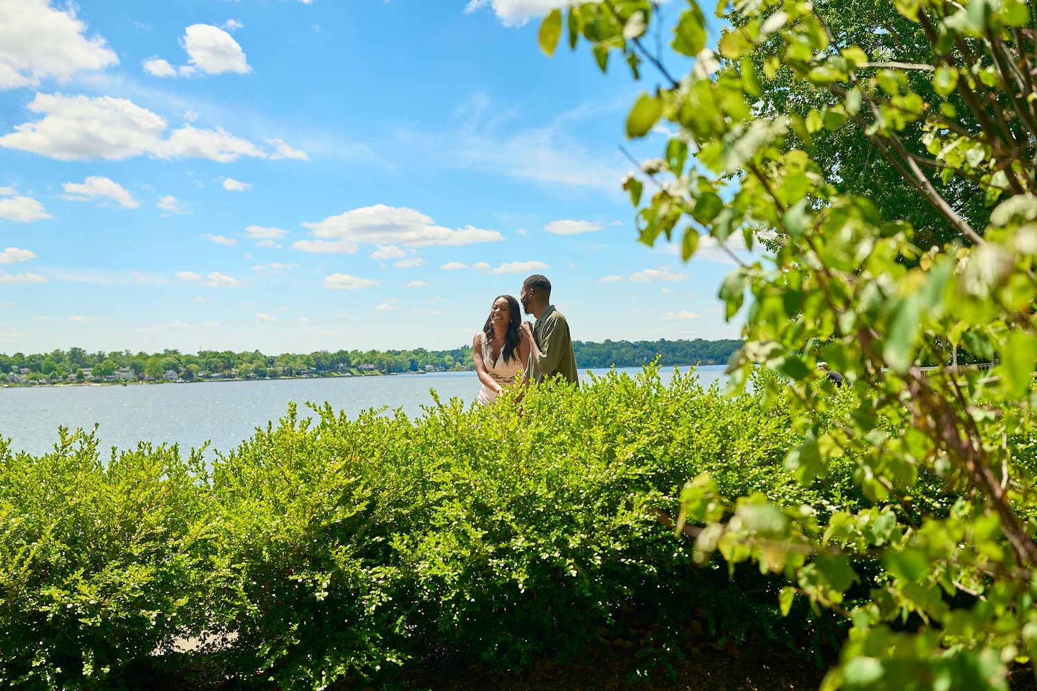 Couple standing together overlooking a serene lake surrounded by lush green bushes under a bright blue sky.
