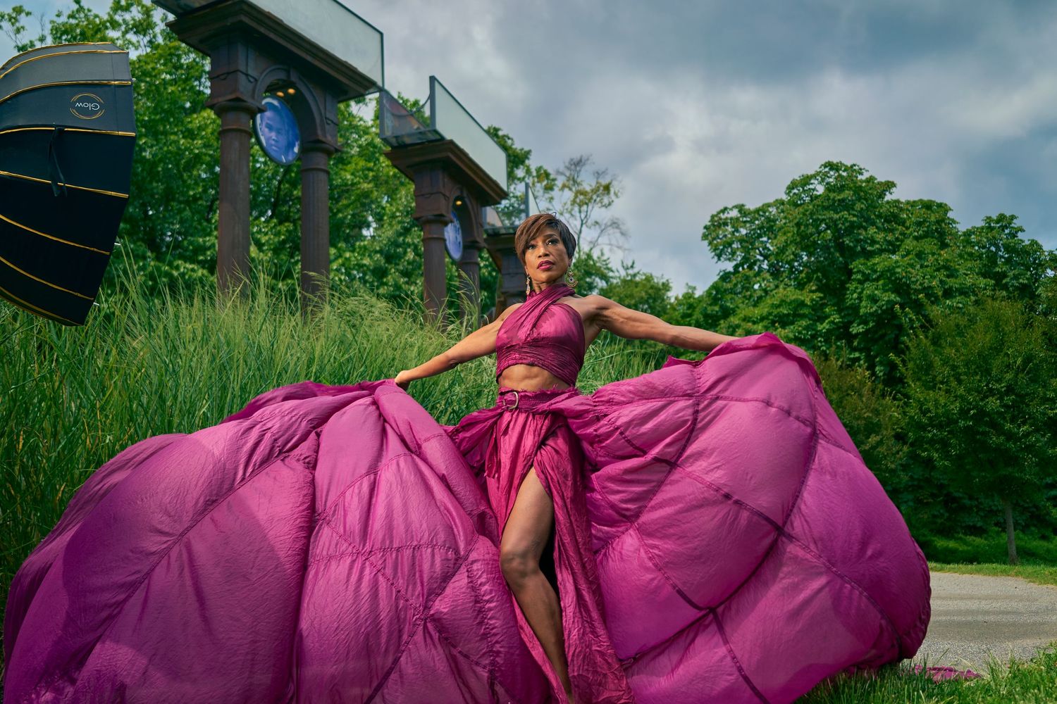 Woman in flowing magenta gown poses outdoors in lush green park with architectural structure in background.