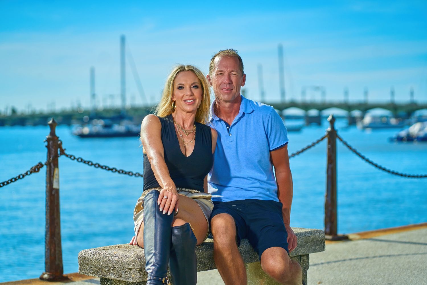 Couple sitting together on waterfront boardwalk with boats and harbor in background.