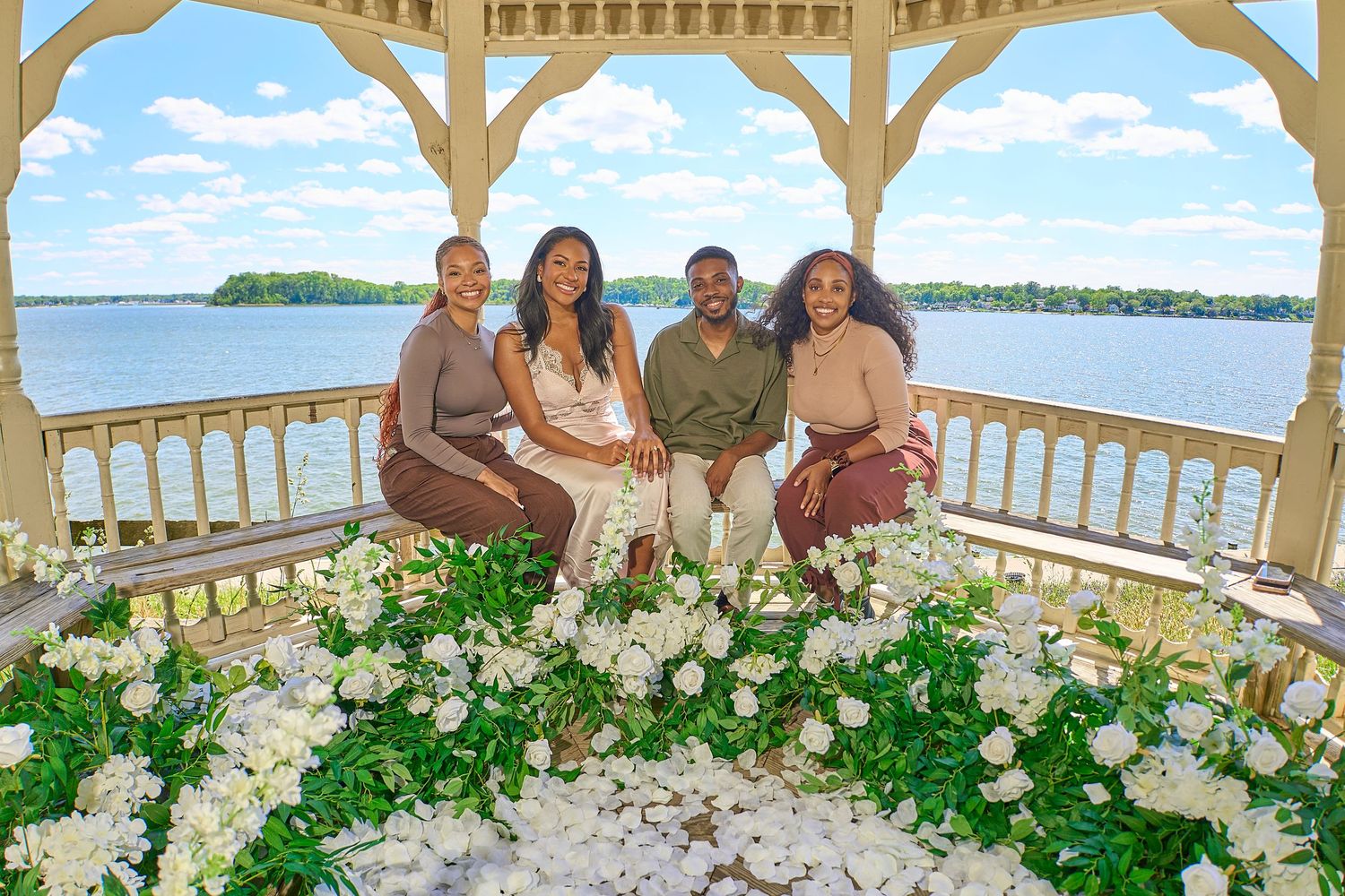 Four people pose in a gazebo surrounded by white flowers with a scenic lake view in the background.