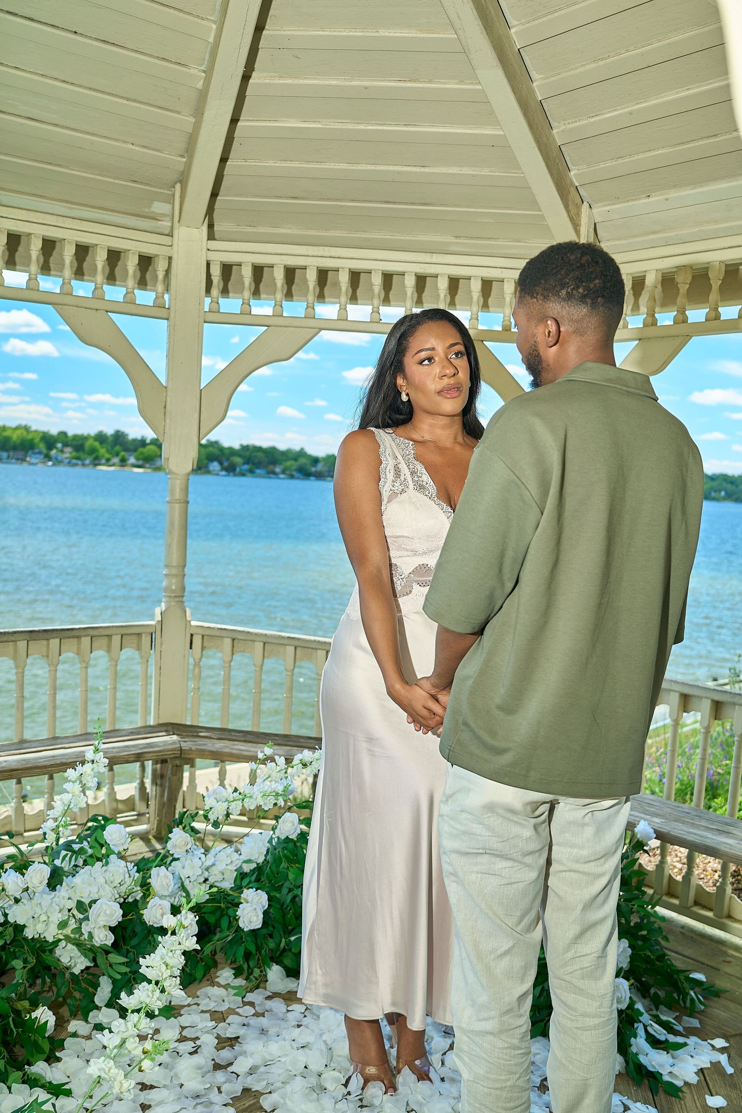 Couple holding hands in a white gazebo decorated with flowers overlooking a serene blue lake.
