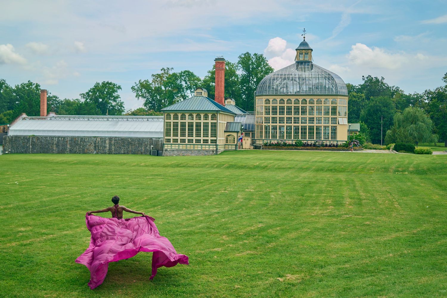 Person in flowing pink fabric poses on green lawn before elegant glass greenhouse conservatory building.