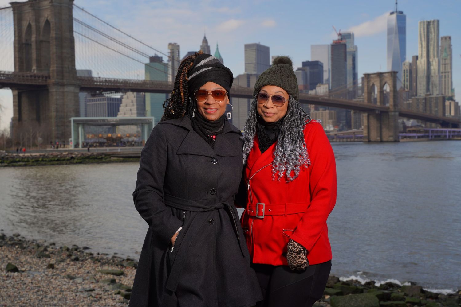 Friends in winter coats taking photos by Brooklyn Bridge with Manhattan skyline view.