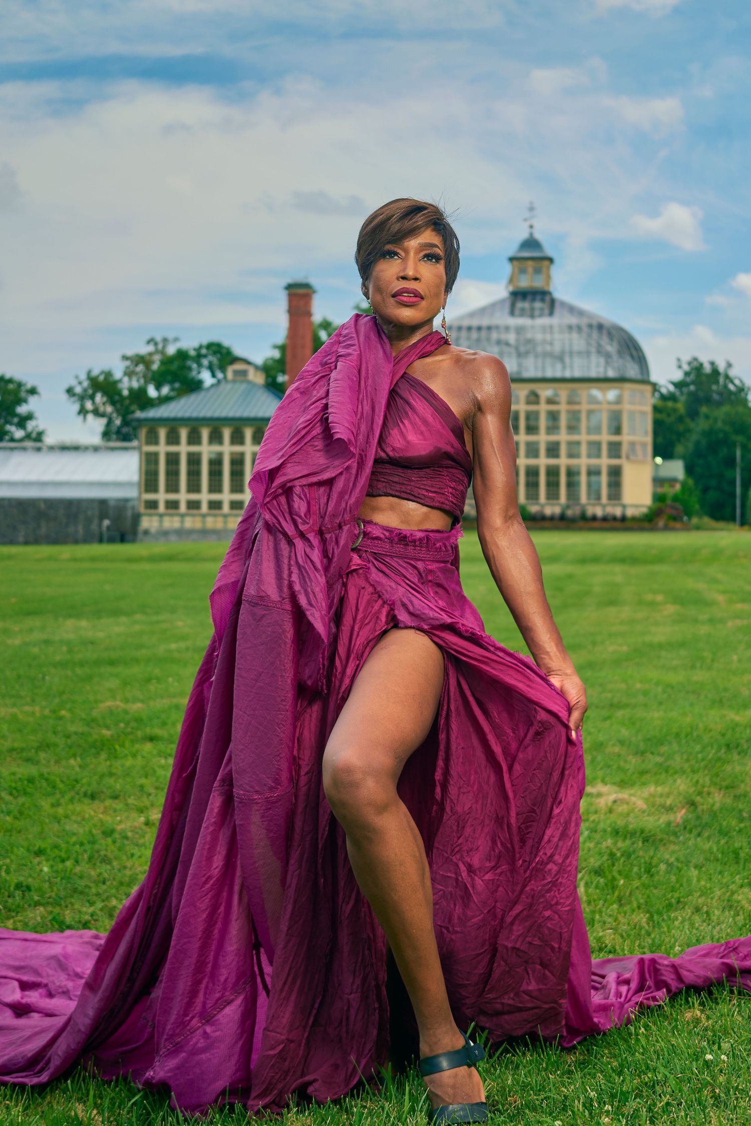 Woman in flowing magenta gown posing on green lawn before historic greenhouse building.