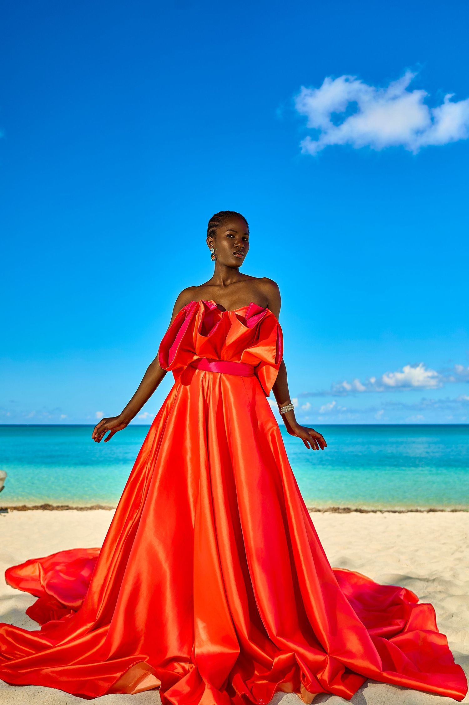 A Black woman in a stunning red satin ballgown poses on a white sand beach with turquoise water and blue sky.