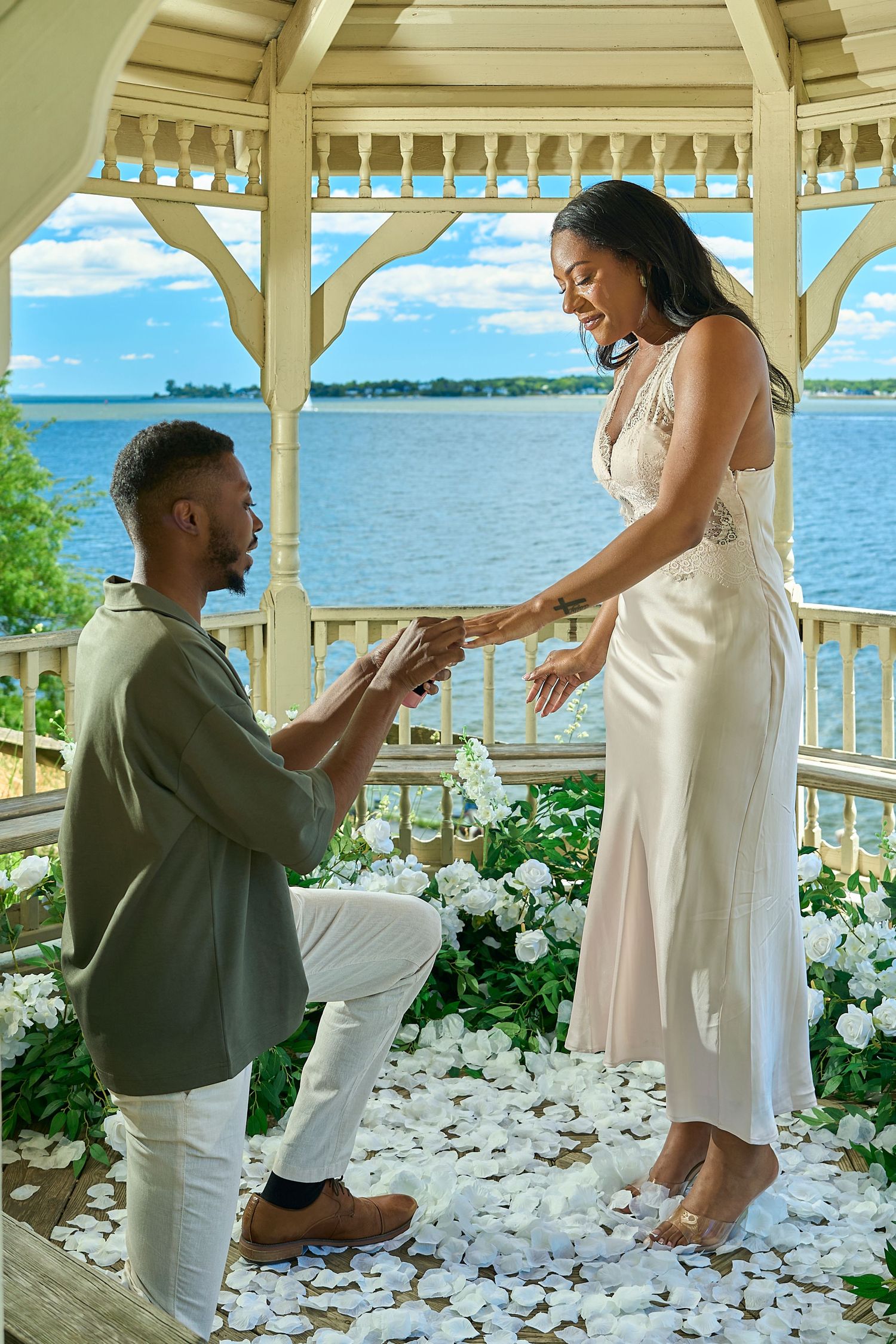 Man proposing on one knee to woman in a white gazebo adorned with white roses and flower petals.