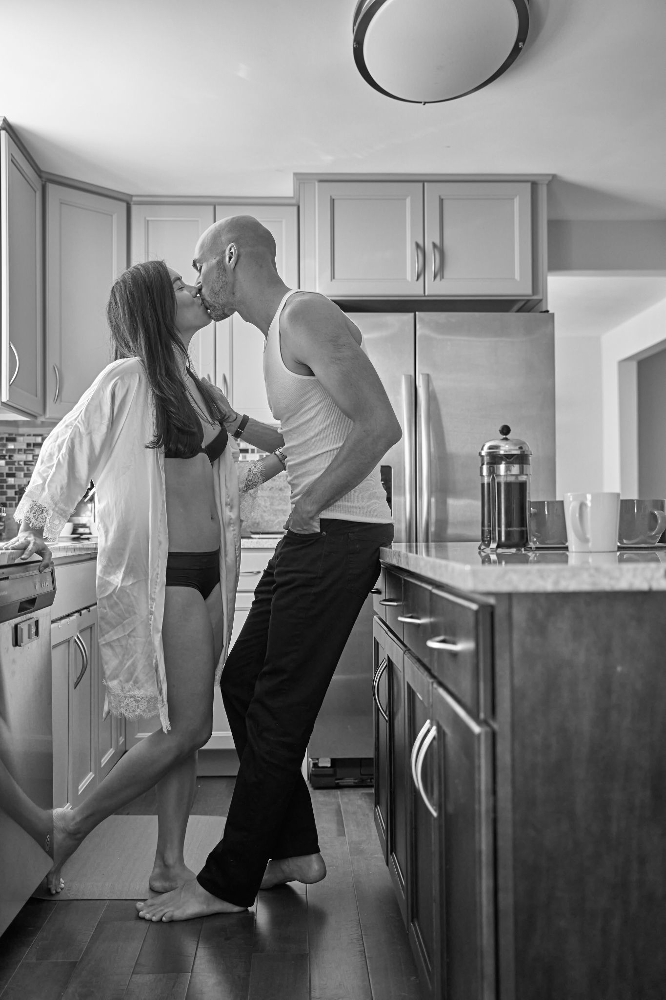 A couple interacts intimately against a kitchen counter in monochrome.