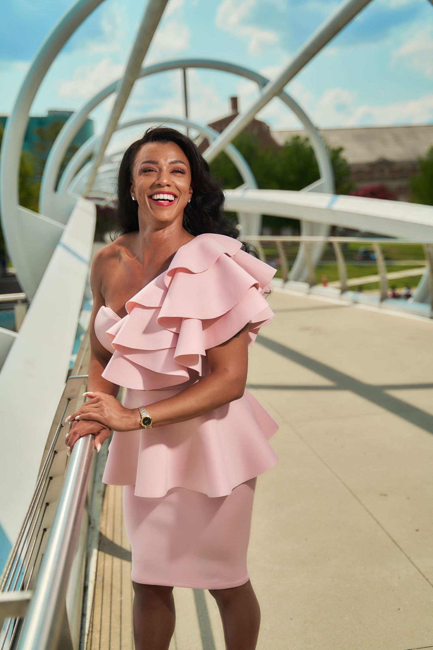 Woman wearing pink ruffled dress smiling and posing on modern white bridge structure.