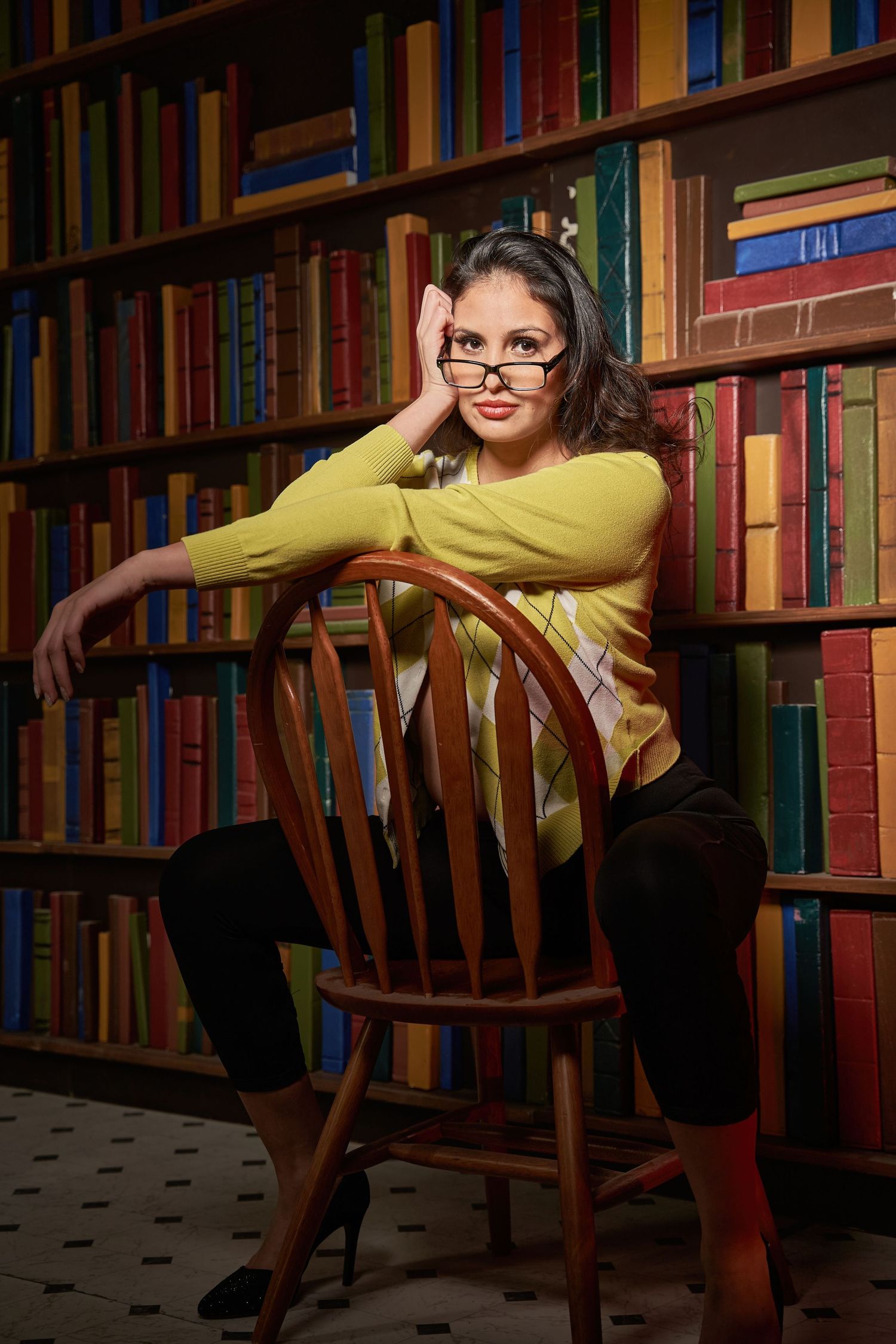 Creative portrait series in library setting with yellow sweater against wooden chair and bookshelves.