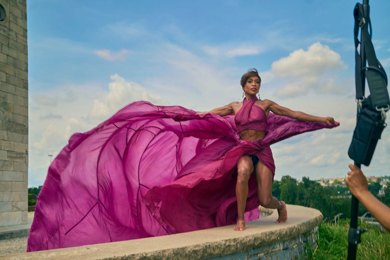 Model in vibrant purple flowing dress poses on stone wall during outdoor fashion photoshoot under blue sky.
