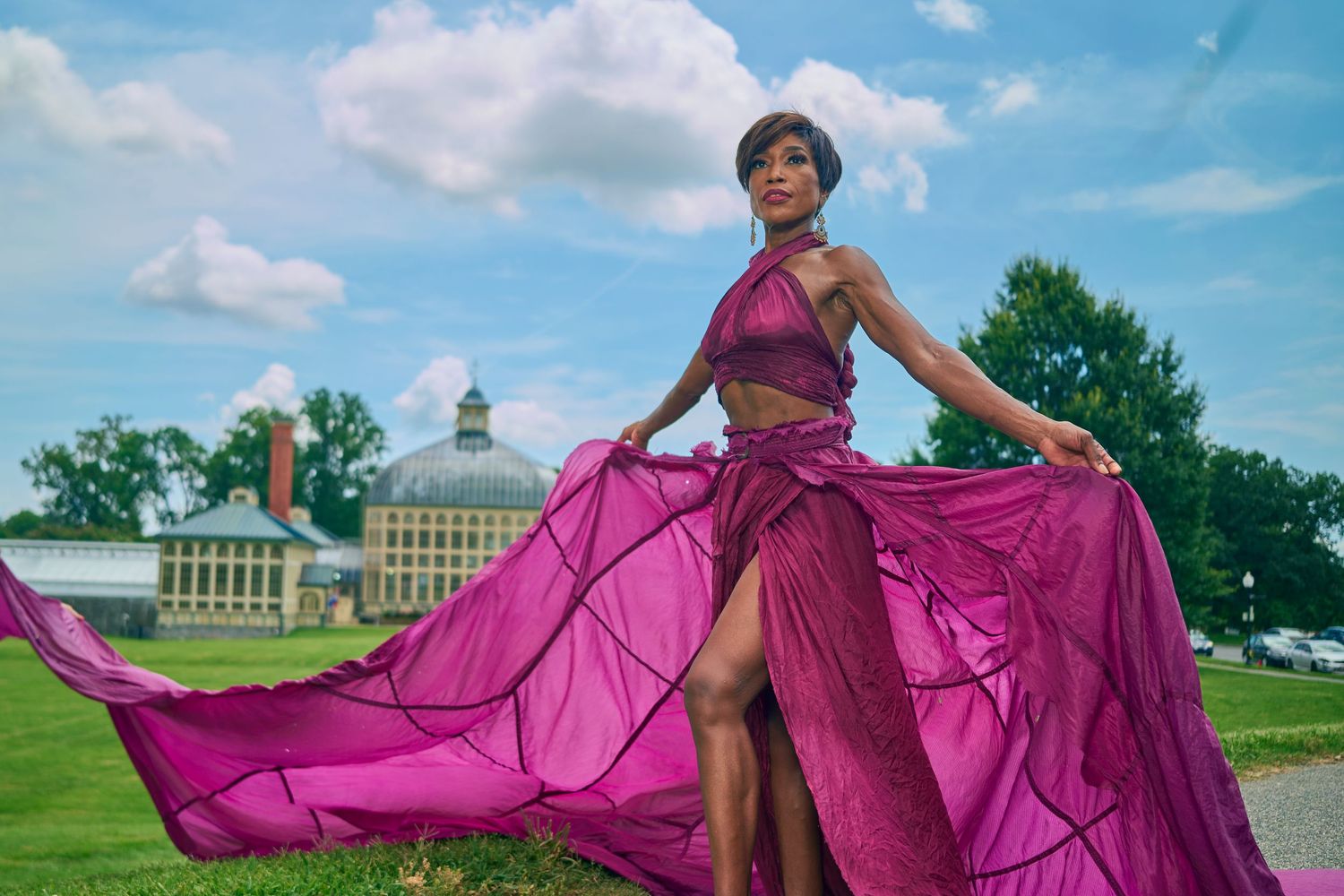 Woman in flowing magenta gown posing outdoors with historic building and cloudy sky in background.