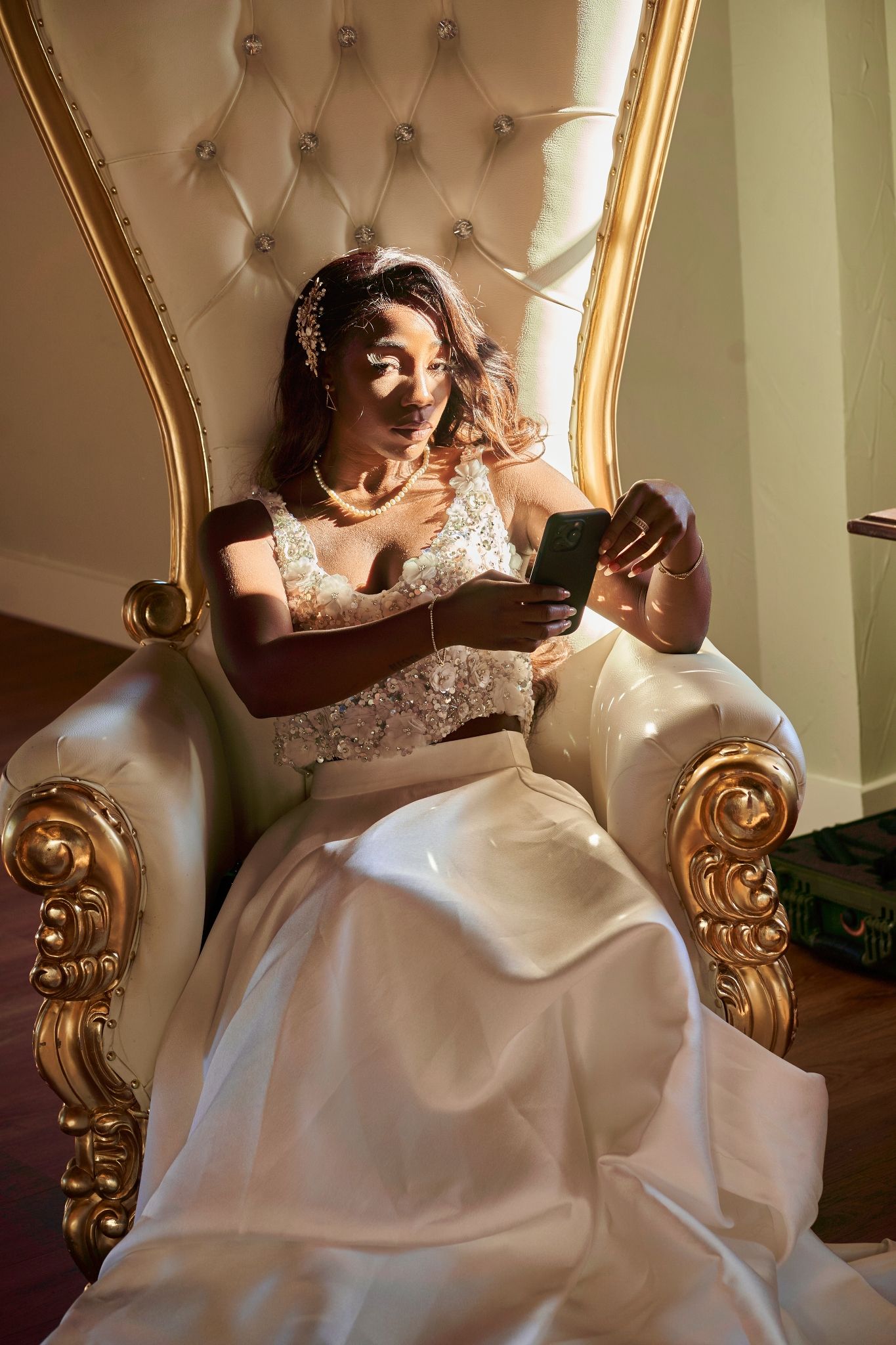 Elegant bride in white gown sitting on ornate gold throne chair checking phone.