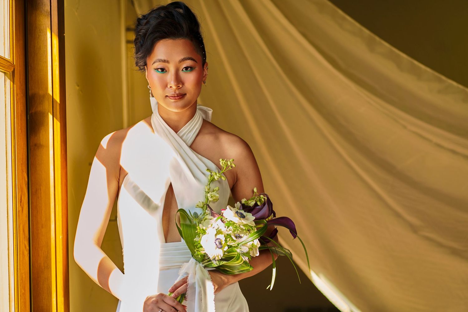 Asian bride in white draped gown holding green and purple floral bouquet standing near golden curtain.