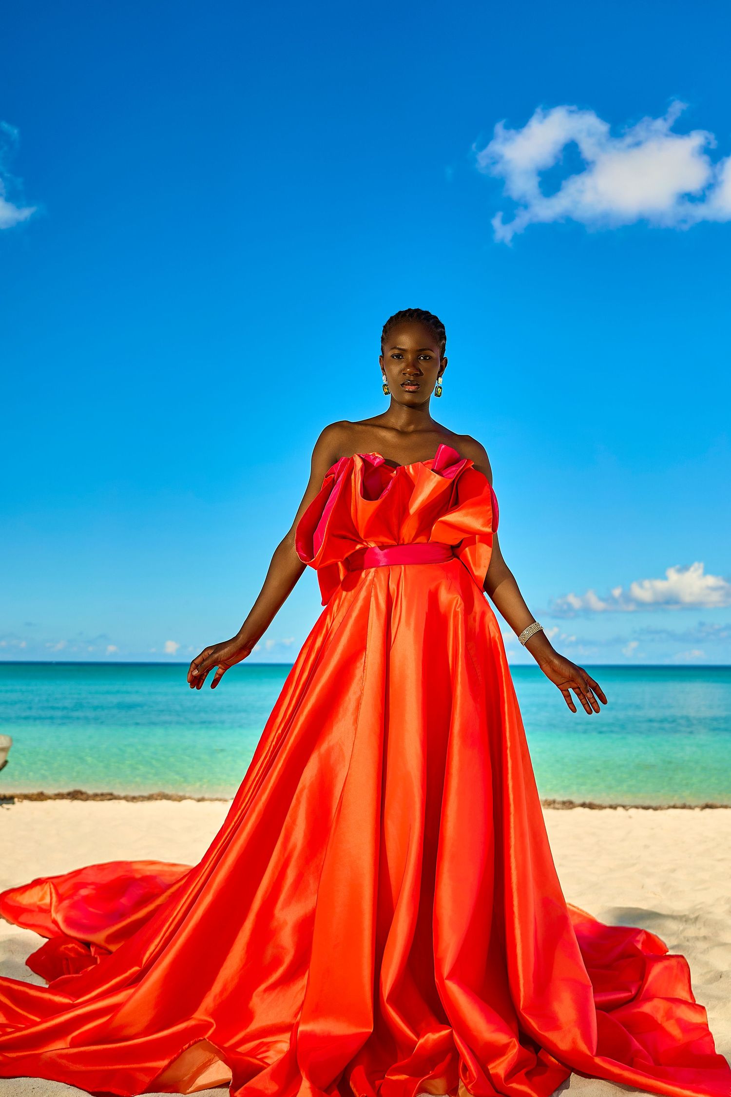 A Black woman in a flowing red-orange satin gown poses on a tropical beach with turquoise water and blue sky.