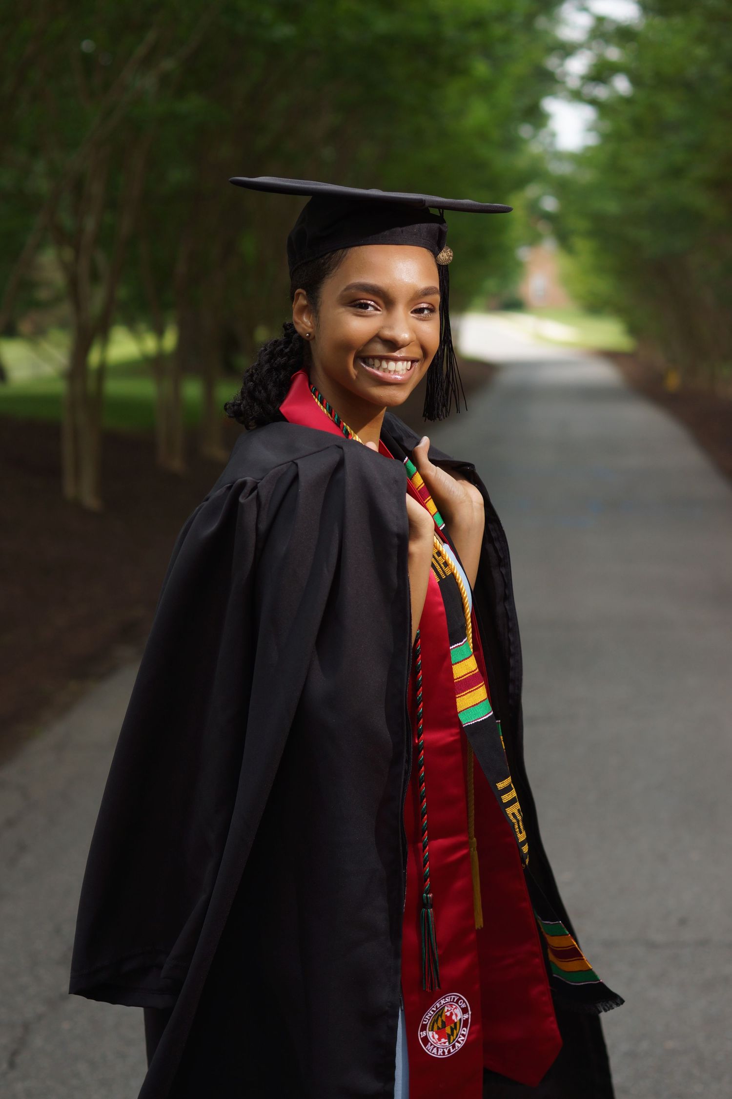Smiling graduate in black cap and gown with colorful stole poses on tree-lined campus pathway.