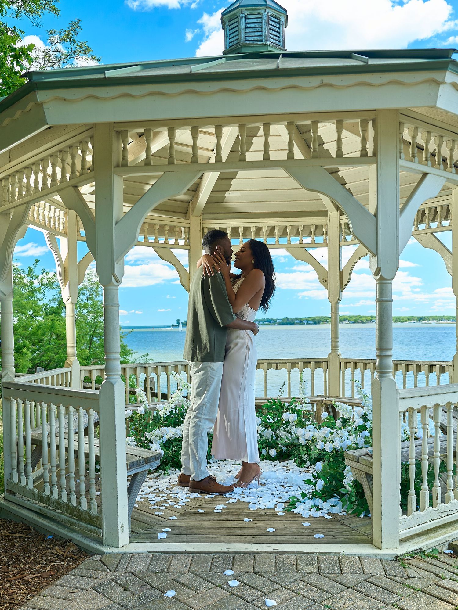 Couple embracing and kissing inside a white gazebo surrounded by white floral arrangements with a scenic lake backdrop.