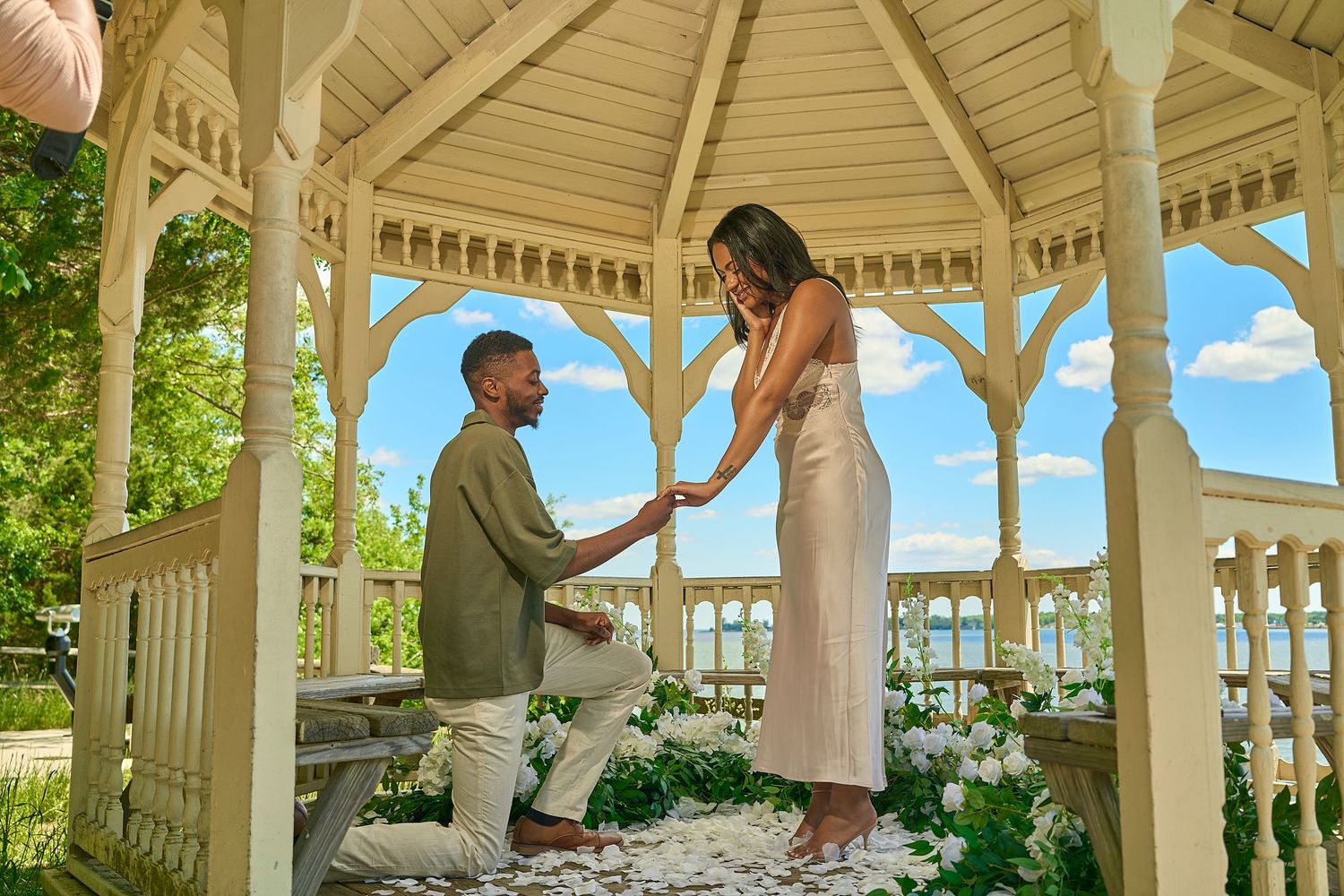 Man proposing to woman in a white gazebo surrounded by flowers on a sunny day.