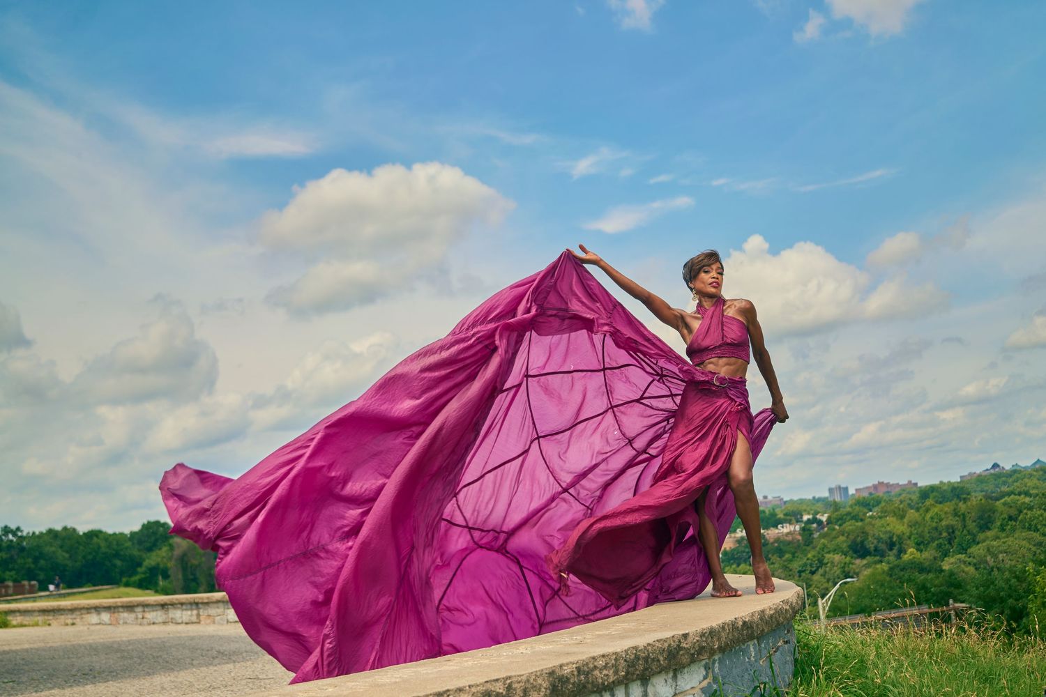 Woman in flowing magenta gown poses on ledge with fabric billowing in wind against blue sky.